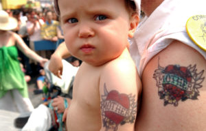 NEW YORK - JUNE 26: A baby sports a "Mother" tattoo like his dad's as he's carried through throngs of onlookers during the 2004 Mermaid Parade June 26, 2004 at Coney Island in the Brooklyn borough of New York. The parade celebrates the sand, the sea and the beginning of summer as well as the mythology of Coney Island. (Photo by Stephen Chernin/Getty Images)